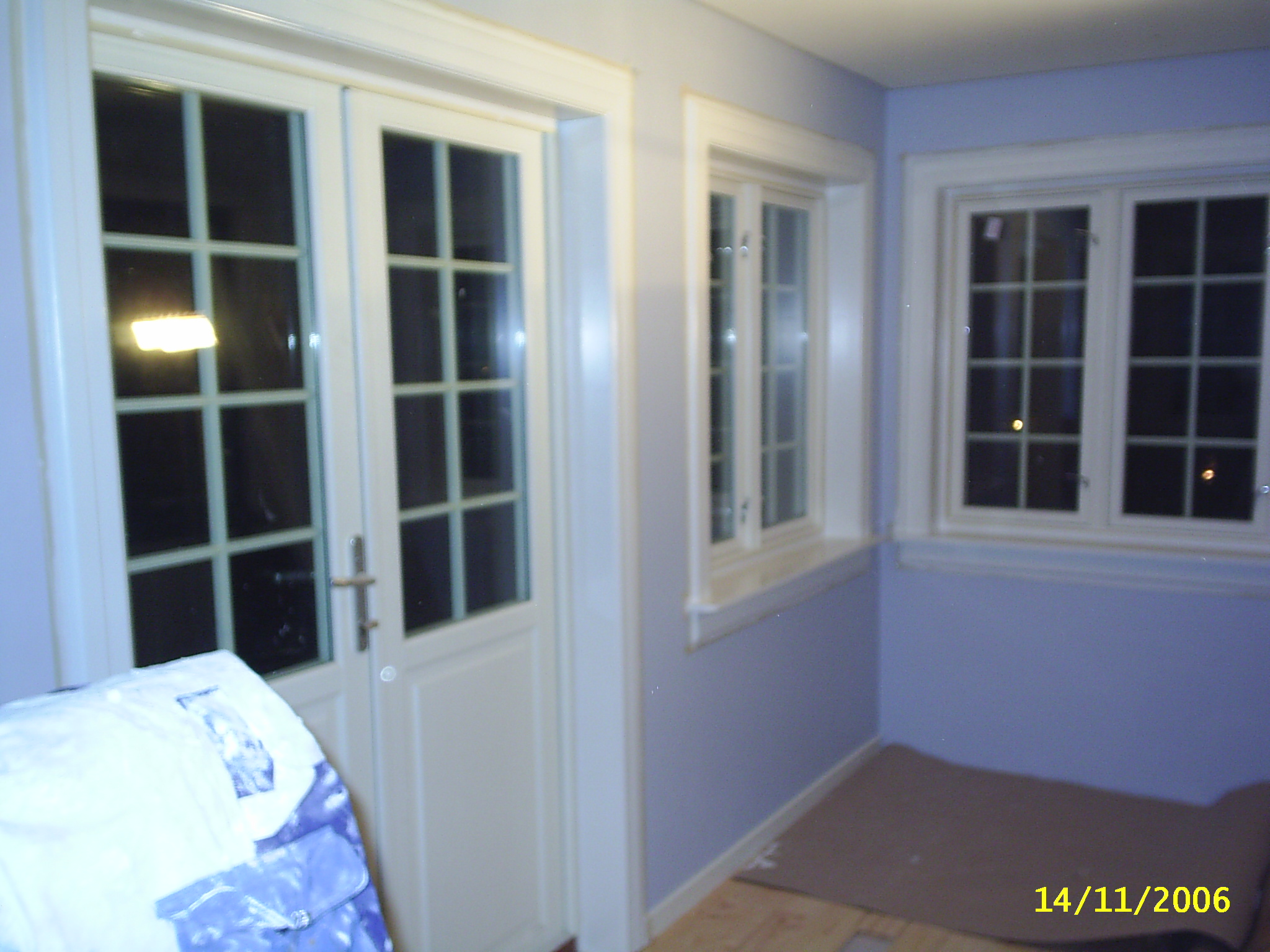 Pokój z pomalowanymi na jasny fiolet ścianami, białymi oknami z kratownicą i podwójnymi drzwiami. Podłoga częściowo zakryta brązowym materiałem ochronnym, w rogu widoczny worek z materiałami...